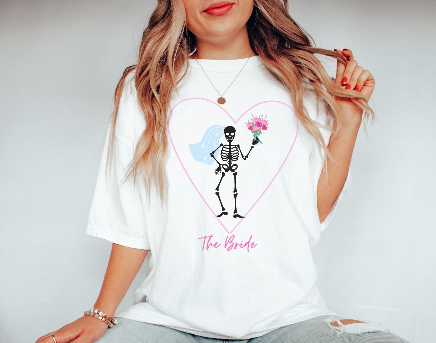 A woman wearing a cute white shirt with a pink heart outline with a black skeleton bride inside wearing a veil and holding a bouquet of flowers with her hand on her hip.