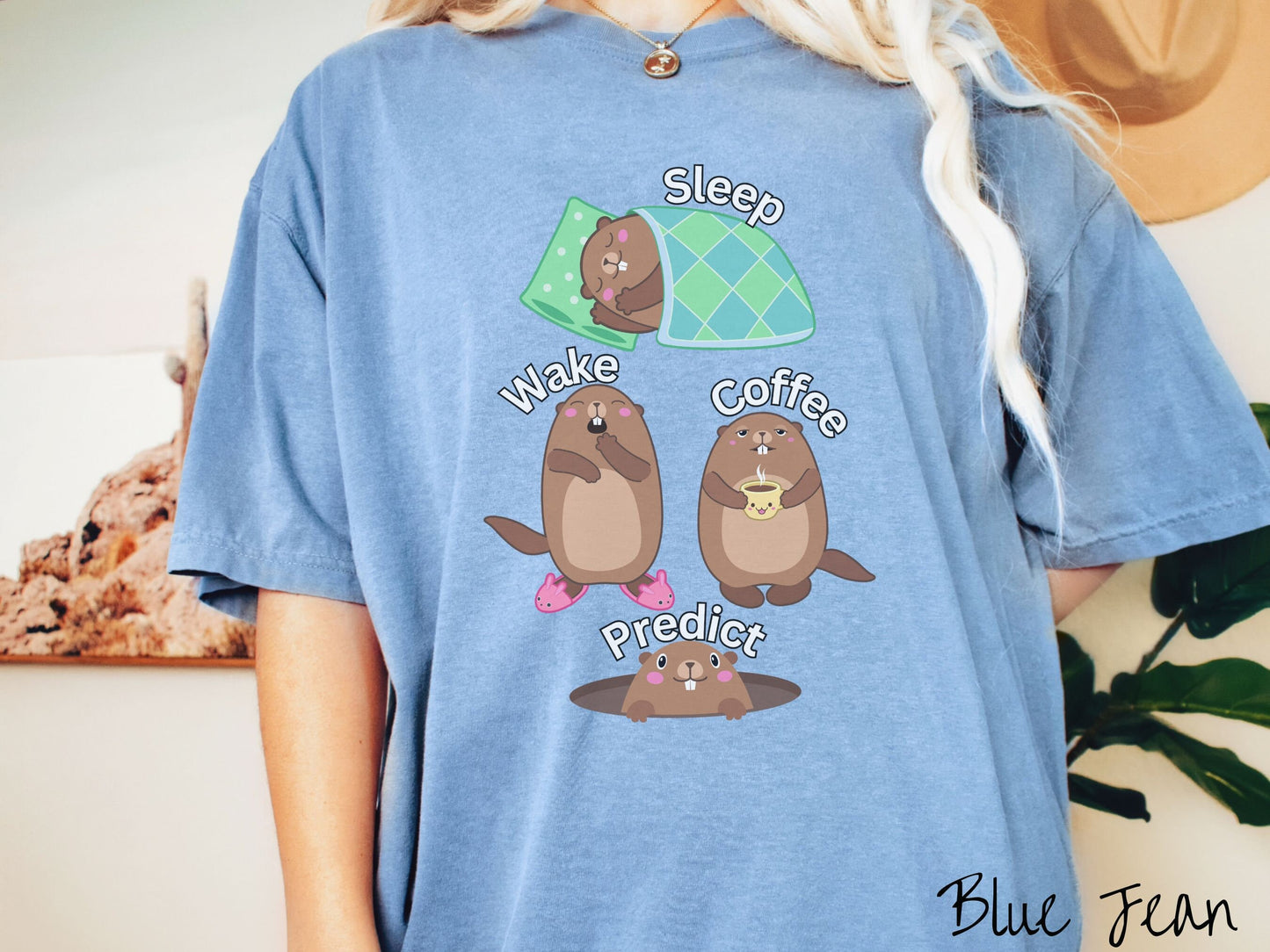 A woman wearing a cute blue jean colored shirt with the text Sleep, Wake, Coffee, and Predict accompanied with pictures of a groundhog sleeping, a groundhog waking up yawning, one holding a steaming cup of coffee, and another exiting its hole.