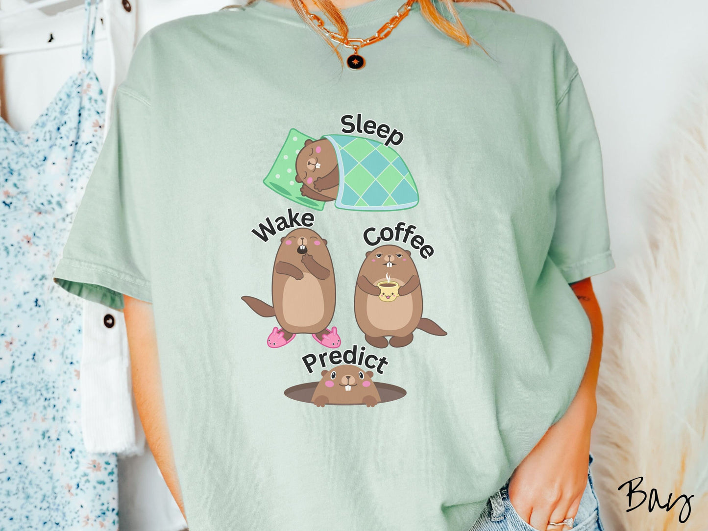 A woman wearing a cute bay colored shirt with the text Sleep, Wake, Coffee, and Predict accompanied with pictures of a groundhog sleeping, a groundhog waking up yawning, one holding a steaming cup of coffee, and another exiting its hole.