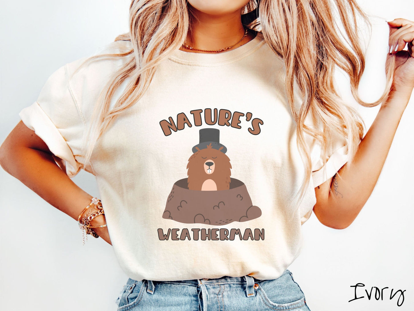 A woman wearing a cute ivory colored shirt with the text Natures Weatherman sandwiching Phil the Punxsutawney PA groundhog who is wearing a black top hat and is coming out of his hole for Groundhogs Day.