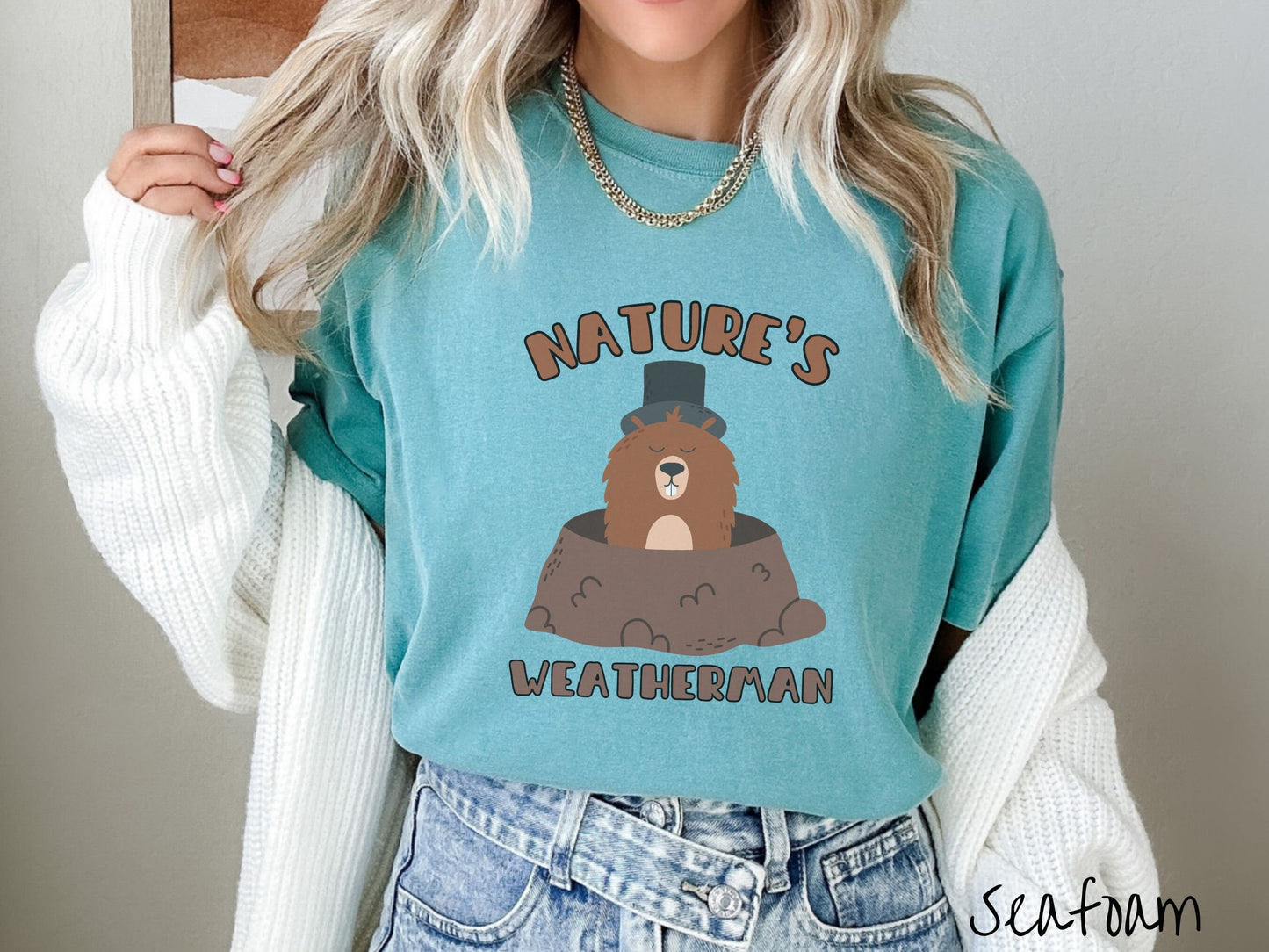 A woman wearing a cute seafoam colored shirt with the text Natures Weatherman sandwiching Phil the Punxsutawney PA groundhog who is wearing a black top hat and is coming out of his hole for Groundhogs Day.