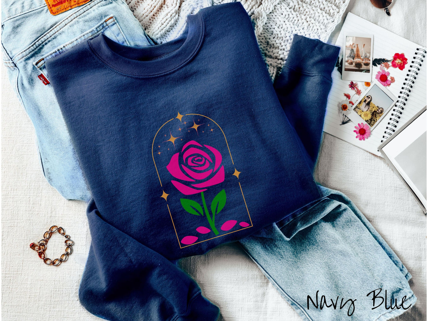 A cute, vintage navy blue colored sweatshirt with an enchanted pink rose in a golden clear container with gold stars twinkling above and pink flower petals resting below.