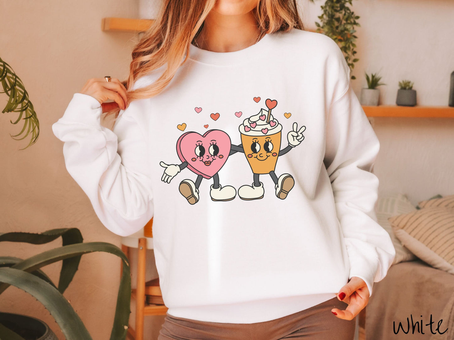 A woman wearing a cute, vintage white colored sweatshirt with colorful hearts above, a female heart with smiling face, legs, and arms wrapping her left arm around a whipped cream topped light brown coffee cup with a heart-tipped topper.
