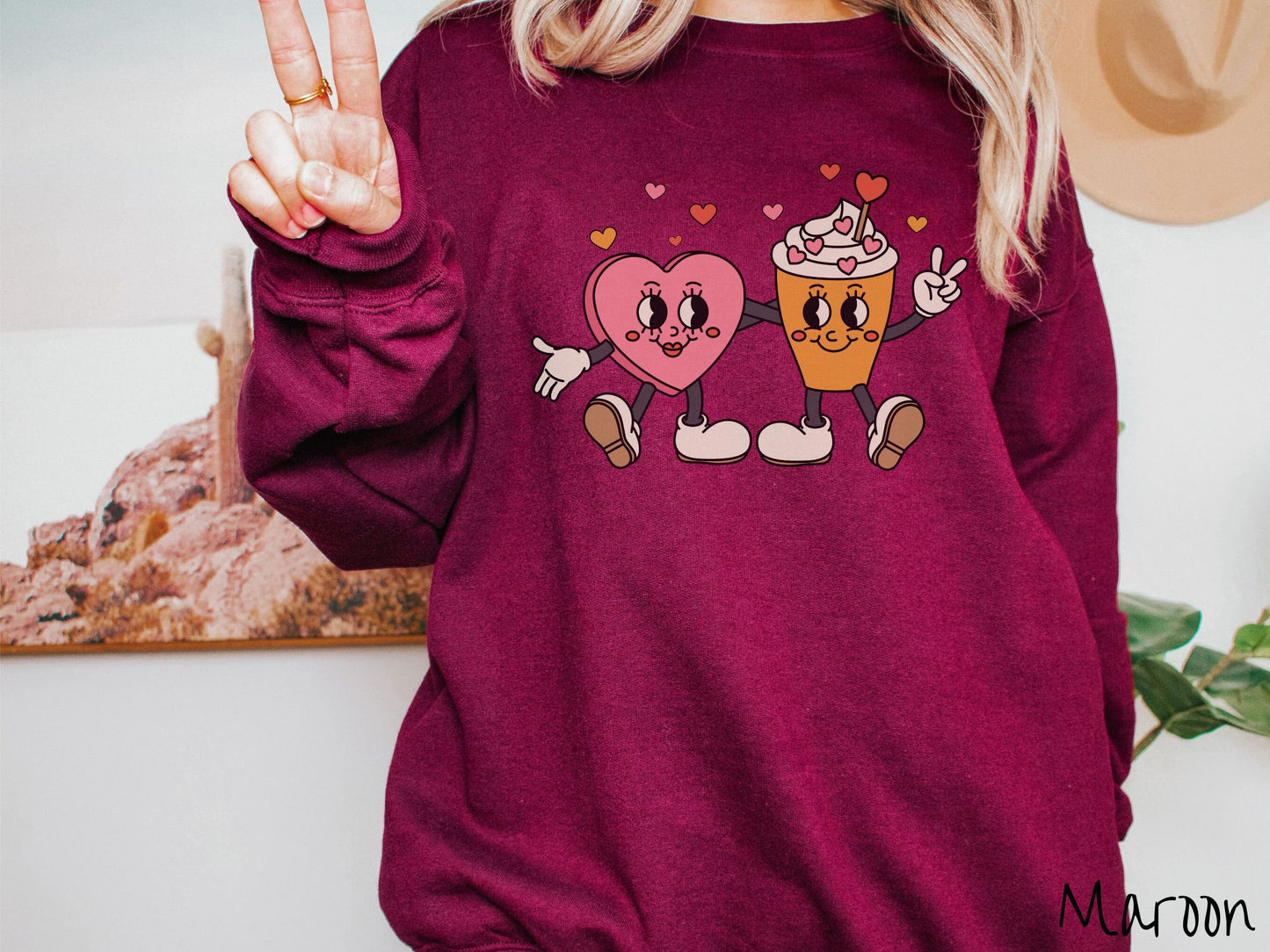 A woman wearing a cute, vintage maroon colored sweatshirt with colorful hearts above, a female heart with smiling face, legs, and arms wrapping her left arm around a whipped cream topped light brown coffee cup with a heart-tipped topper.