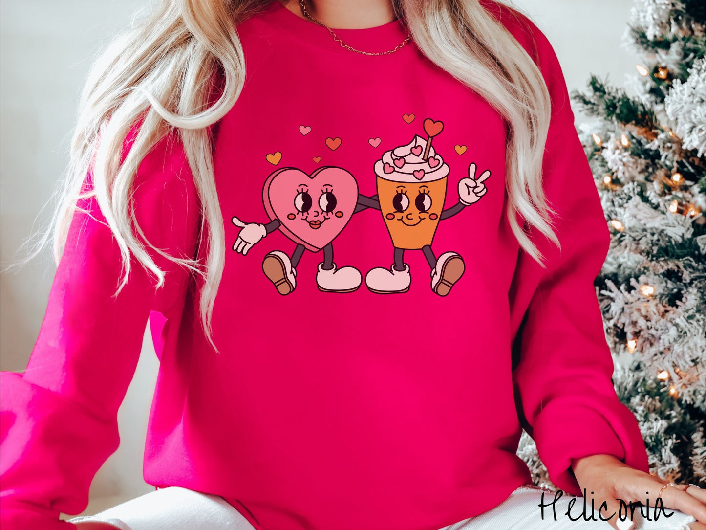 A woman wearing a cute, vintage heliconia colored sweatshirt with colorful hearts above, a female heart with smiling face, legs, and arms wrapping her left arm around a whipped cream topped light brown coffee cup with a heart-tipped topper.