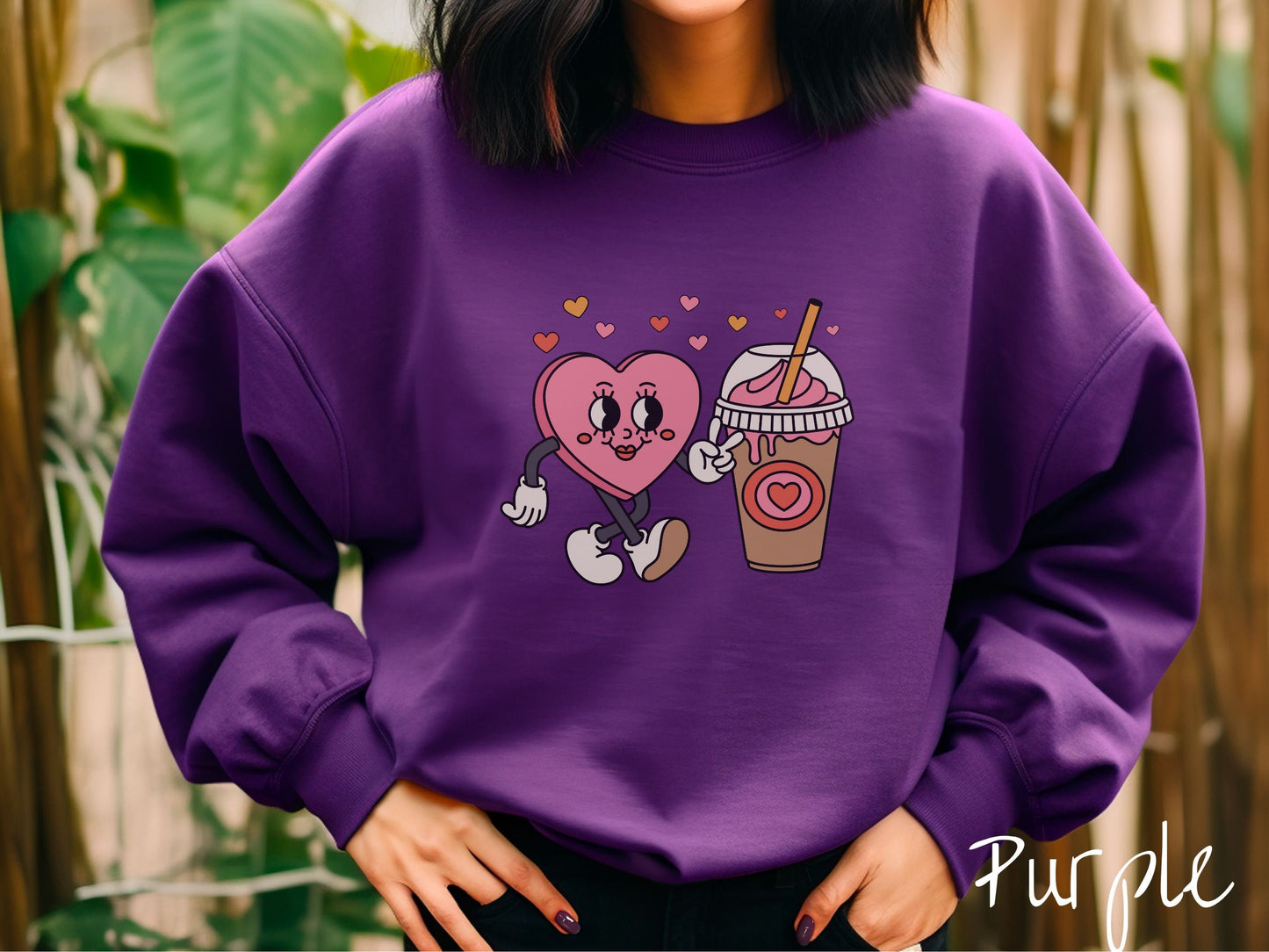 A woman wearing a cute, vintage purple colored sweatshirt and colorful hearts above, a female heart with smiling face, legs, and arms showing a peace sign with her left hand next to an iced coffee and pink whipped cream and red heart logo on the cup.