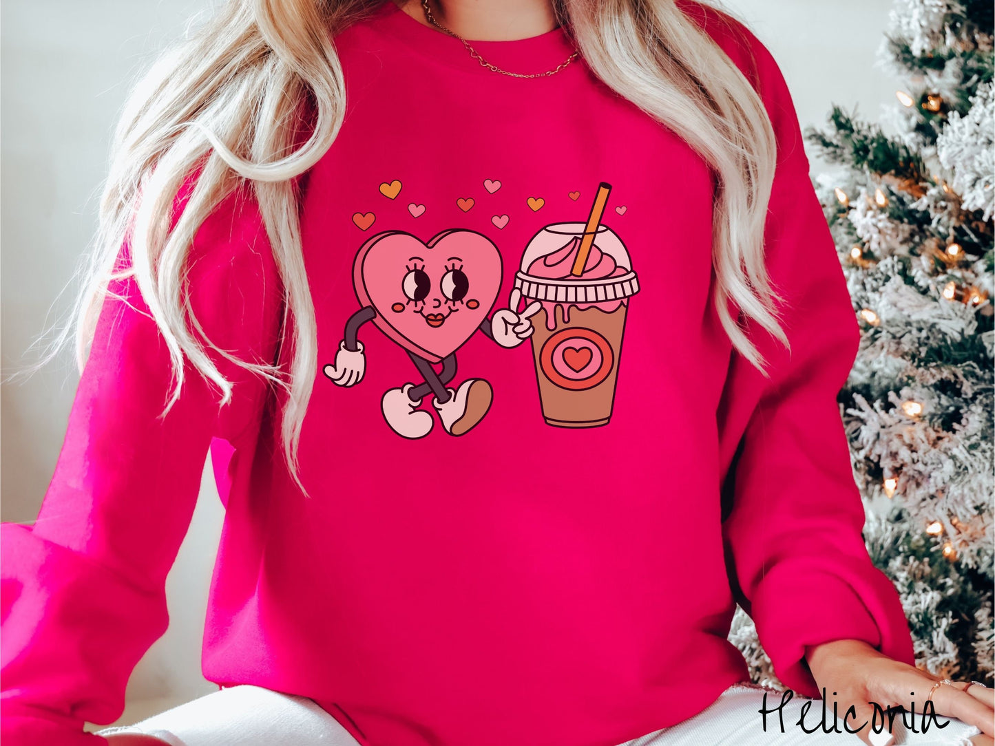 A woman wearing a cute, vintage heliconia colored sweatshirt and hearts above, a female heart with smiling face, legs, and arms showing a peace sign with her left hand next to an iced coffee and pink whipped cream and a red heart logo on the cup.