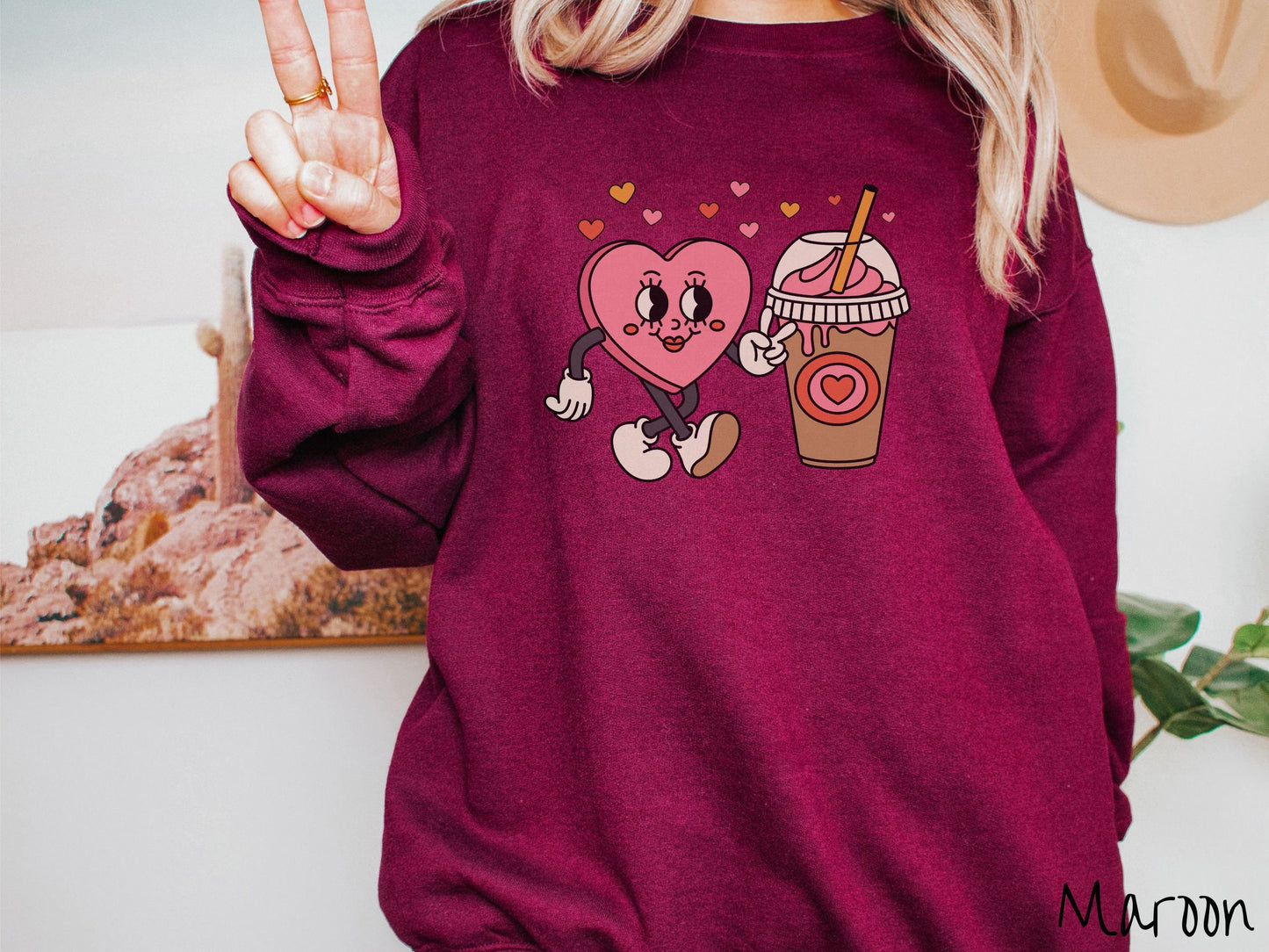 A woman wearing a cute, vintage maroon colored sweatshirt and hearts above, a female heart with smiling face, legs, and arms showing a peace sign with her left hand next to an iced coffee and pink whipped cream and a red heart logo on the cup.