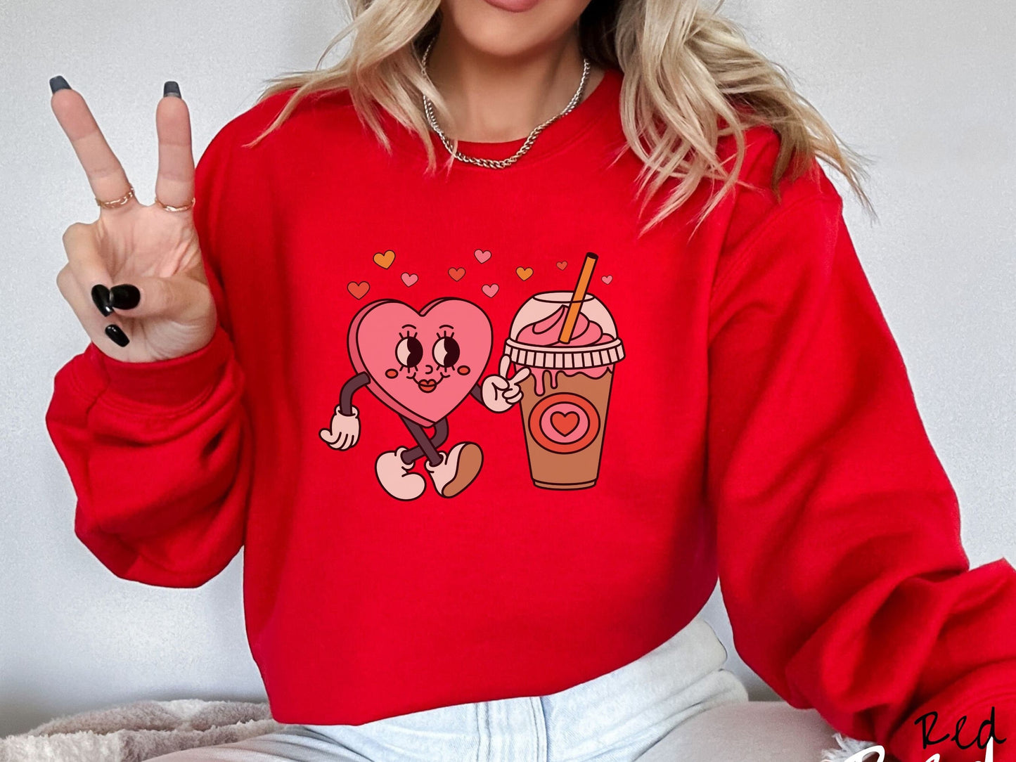 A woman wearing a cute, vintage red colored sweatshirt with colorful hearts above, a female heart with smiling face, legs, and arms showing a peace sign with her left hand next to an iced coffee and pink whipped cream and a red heart logo on the cup.