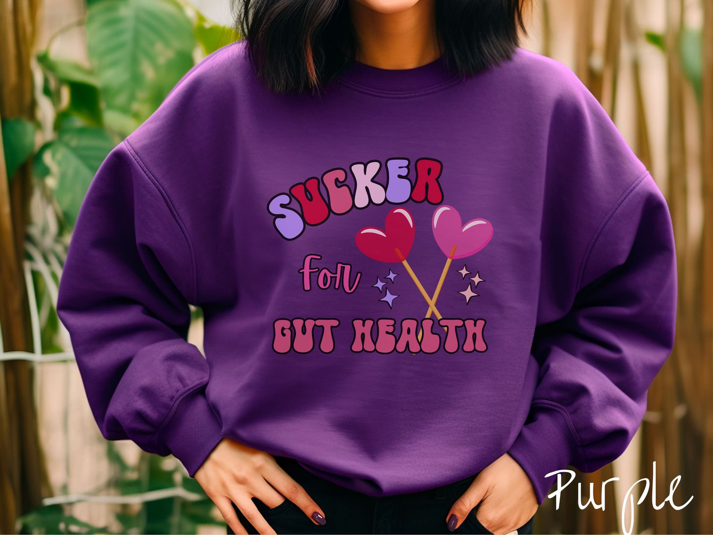 A woman wearing a cute, vintage purple colored sweatshirt with the text Sucker for Gut Health in red, pink, and purple, and there are two lollipops with pink and red hearts on the ends of the sticks and purple and yellow stars in the background.