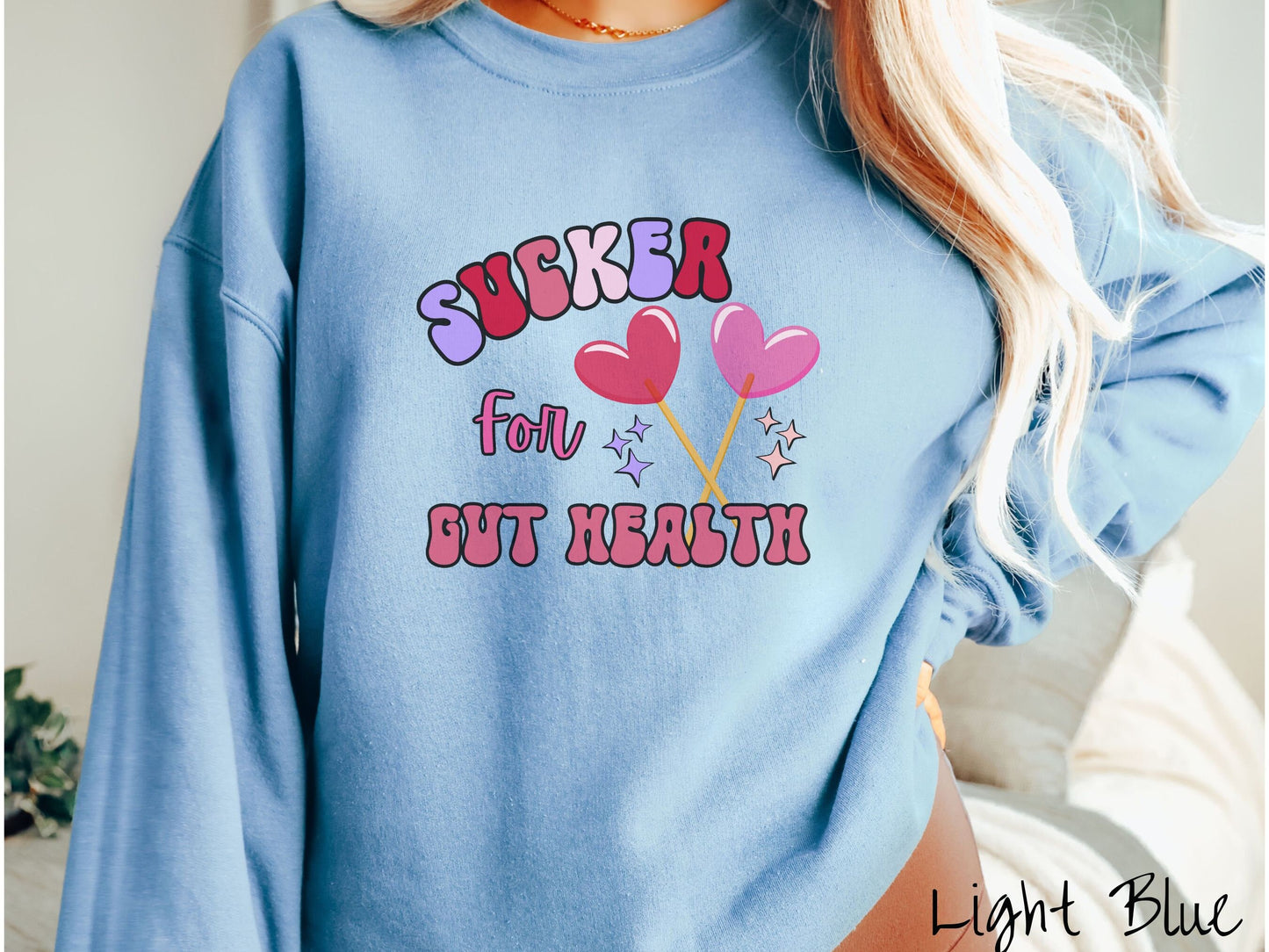 A woman wearing a cute, vintage light blue colored sweatshirt with the text Sucker for Gut Health in red, pink, and purple, and there are two lollipops with pink and red hearts on the ends of the sticks and purple and yellow stars in the background.