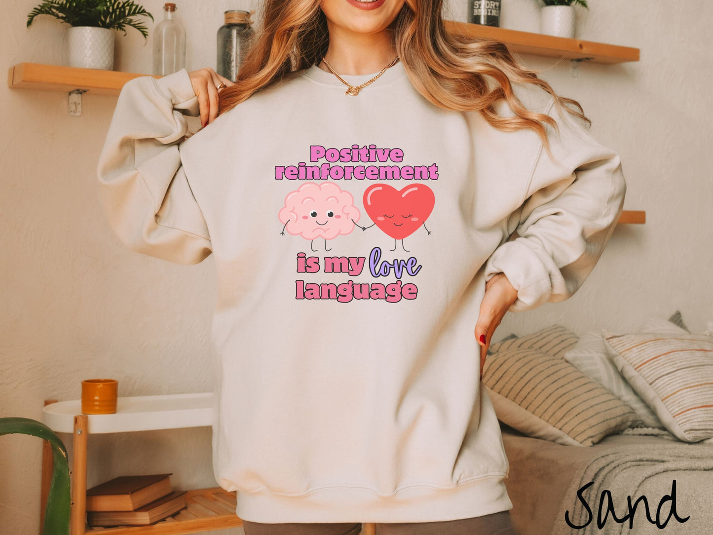 A woman wearing a cute, vintage sand colored sweatshirt with the text Positive reinforcement is my love language in pink, purple, and red with a pink smiling brain holding hands with a red smiling heart.