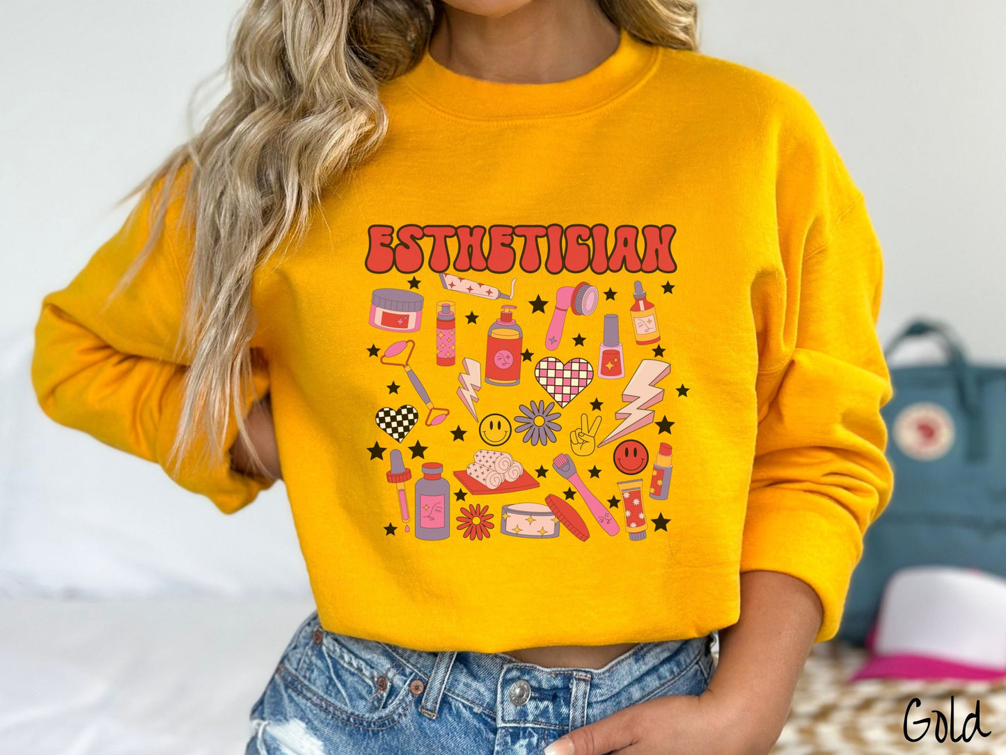 A woman wearing a cute gold colored sweatshirt with the word Esthetician across the top and hair care items below like cream, rollers, hair care products, and droppers. Smiley faces, stars, and lightning bolts are spread out among the items.