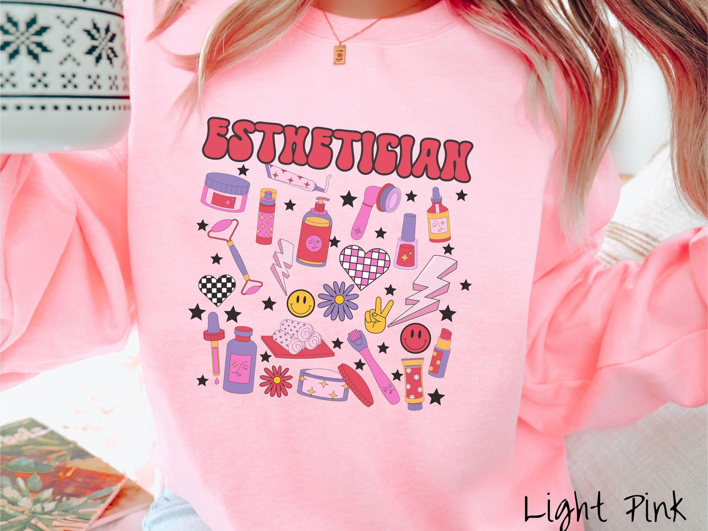 A woman wearing a cute light pink colored sweatshirt with the word Esthetician across the top and hair care items below like cream, rollers, hair care products, and droppers. Smiley faces, stars, and lightning bolts are spread out among the items.