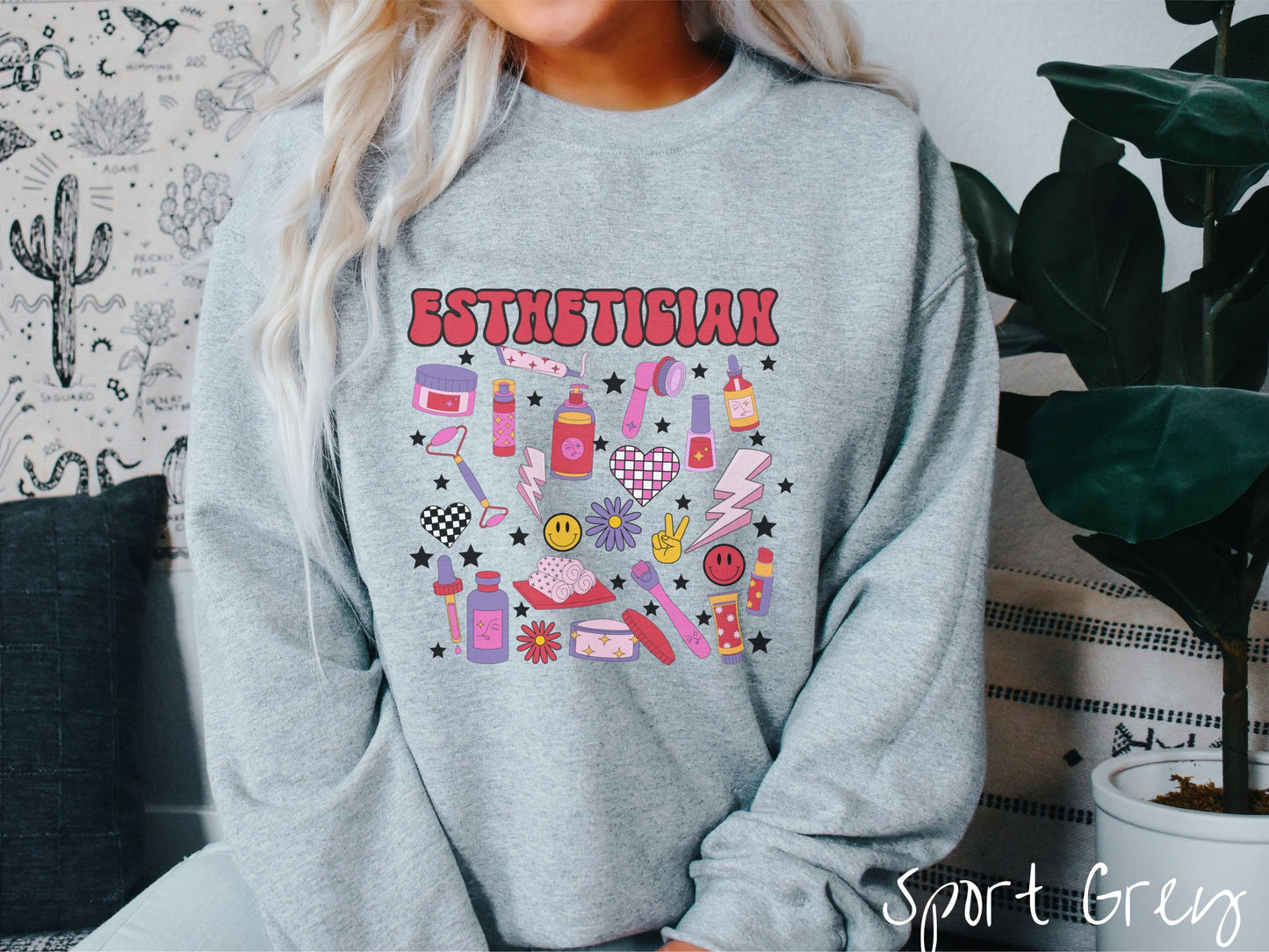 A woman wearing a cute sport grey colored sweatshirt with the word Esthetician across the top and hair care items below like cream, rollers, hair care products, and droppers. Smiley faces, stars, and lightning bolts are spread out among the items.