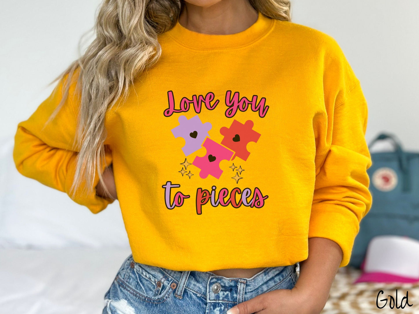 A woman wearing a cute, vintage gold colored sweatshirt with the text Love you to pieces in red, pink, and purple colors, and three red, pink, and purple puzzle pieces with hearts on them surrounded by yellow stars.