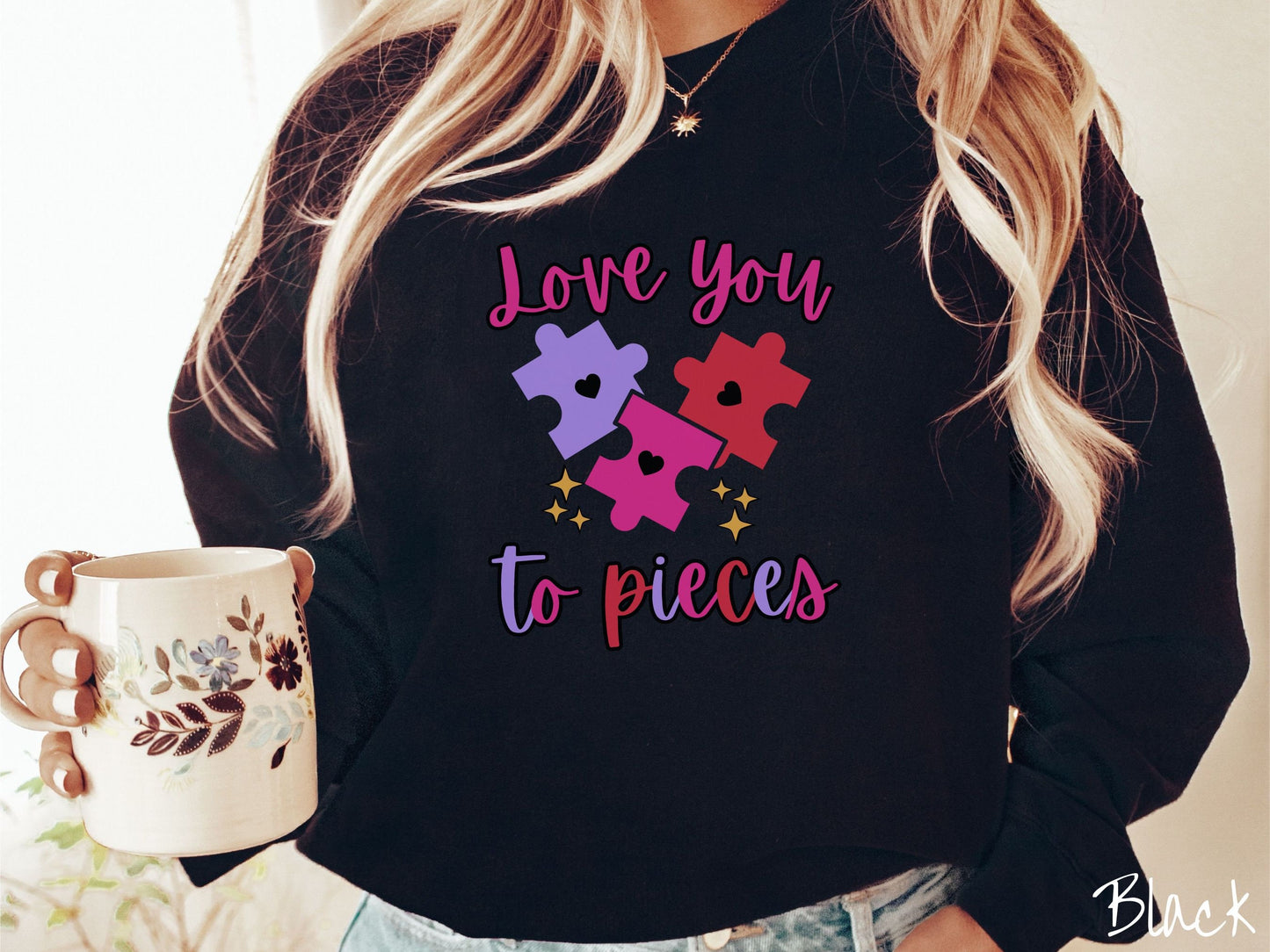 A woman wearing a cute, vintage black colored sweatshirt with the text Love you to pieces in red, pink, and purple colors, and three red, pink, and purple puzzle pieces with hearts on them surrounded by yellow stars.