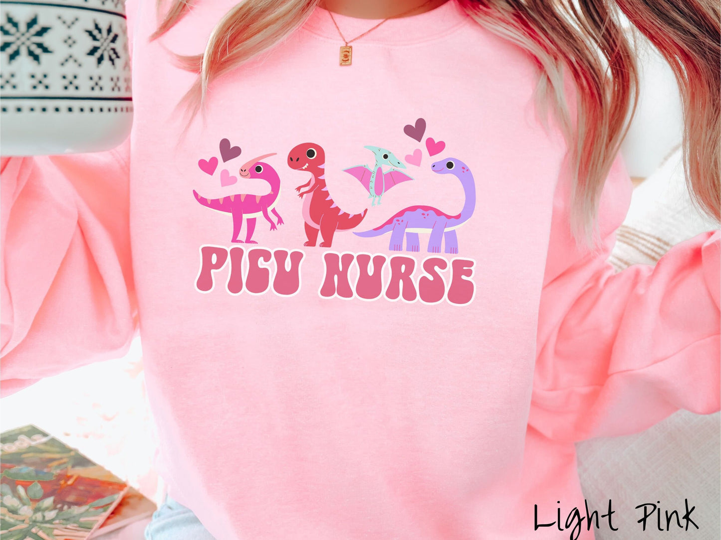 A woman wearing a cute light pink colored sweatshirt with the text PICU Nurse in the center below four dinosaurs, a pink Parasaurolophus, a red tyrannosaurus rex, a green pterodactyl, a purple Brachiosaurus, and pink and red hearts in the air.