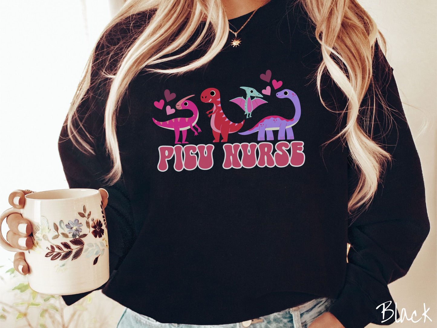 A woman wearing a cute black colored sweatshirt with the text PICU Nurse in the center below four dinosaurs, a pink Parasaurolophus, a red tyrannosaurus rex, a green pterodactyl, a purple Brachiosaurus, and pink and red hearts in the air.