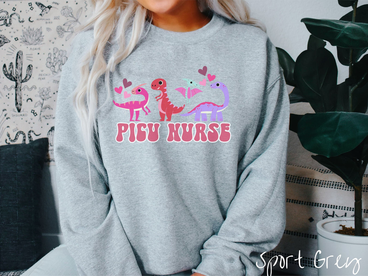 A woman wearing a cute sport grey colored sweatshirt with the text PICU Nurse in the center below four dinosaurs, a pink Parasaurolophus, a red tyrannosaurus rex, a green pterodactyl, a purple Brachiosaurus, and pink and red hearts in the air.