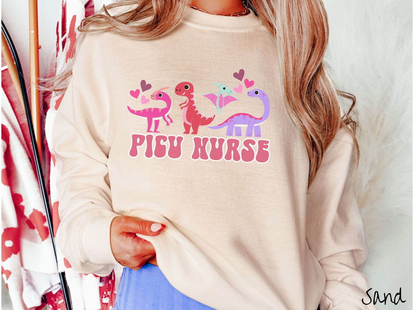 A woman wearing a cute sand colored sweatshirt with the text PICU Nurse in the center below four dinosaurs, a pink Parasaurolophus, a red tyrannosaurus rex, a green pterodactyl, a purple Brachiosaurus, and pink and red hearts in the air.