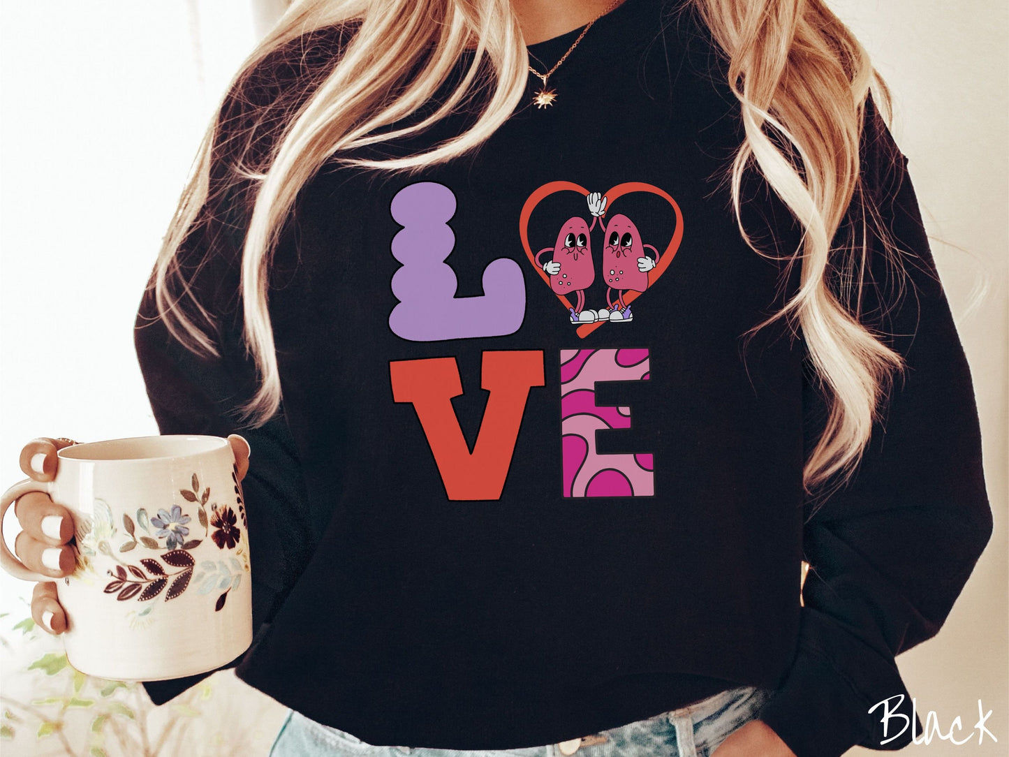 A woman wearing a cute black colored sweatshirt with the word LOVE spelled out with the L as a purple lung, the O as a heart with two lung figures inside high fiving, the V is orange, and the E is two shades of pink.