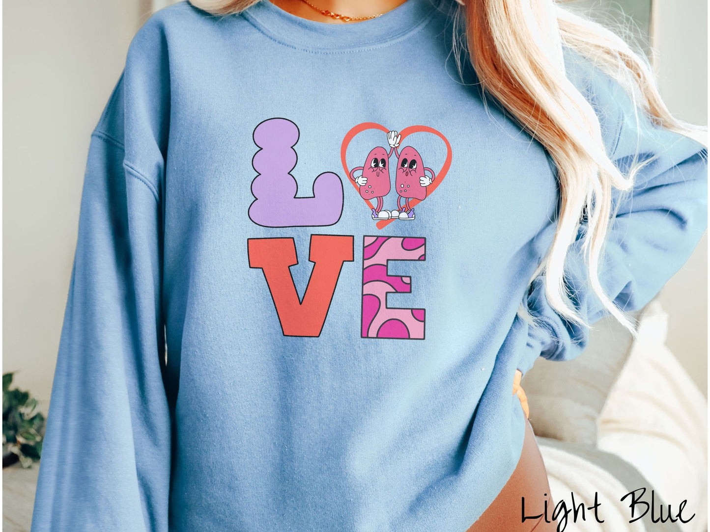 A woman wearing a cute light blue colored sweatshirt with the word LOVE spelled out with the L as a purple lung, the O as a heart with two lung figures inside high fiving, the V is orange, and the E is two shades of pink.