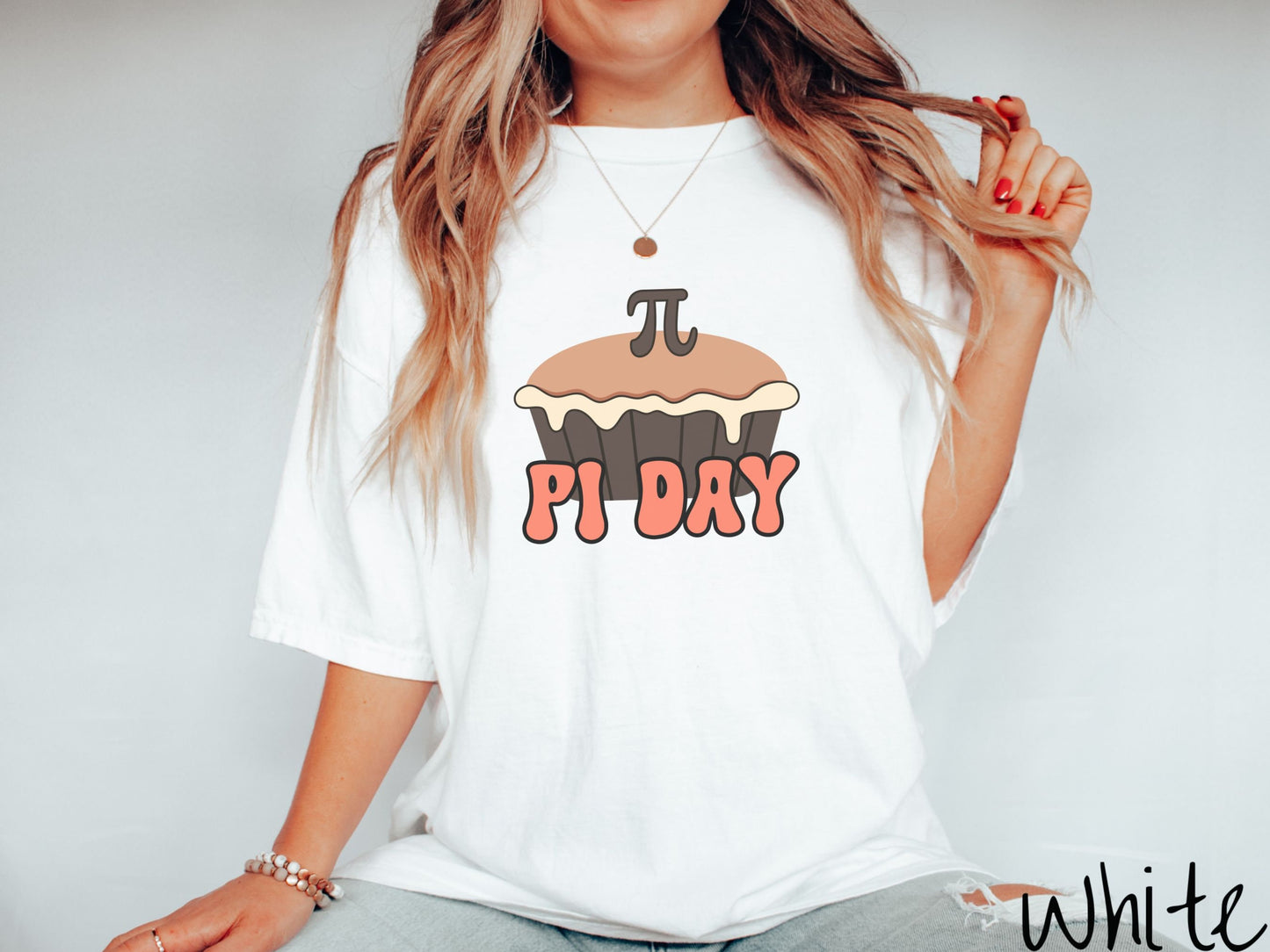 A woman wearing a cute, vintage white colored comfort colors shirt with the text Pi Day in orange font, above that is a brown pumpkin pie with a pi symbol on the top of the pie.