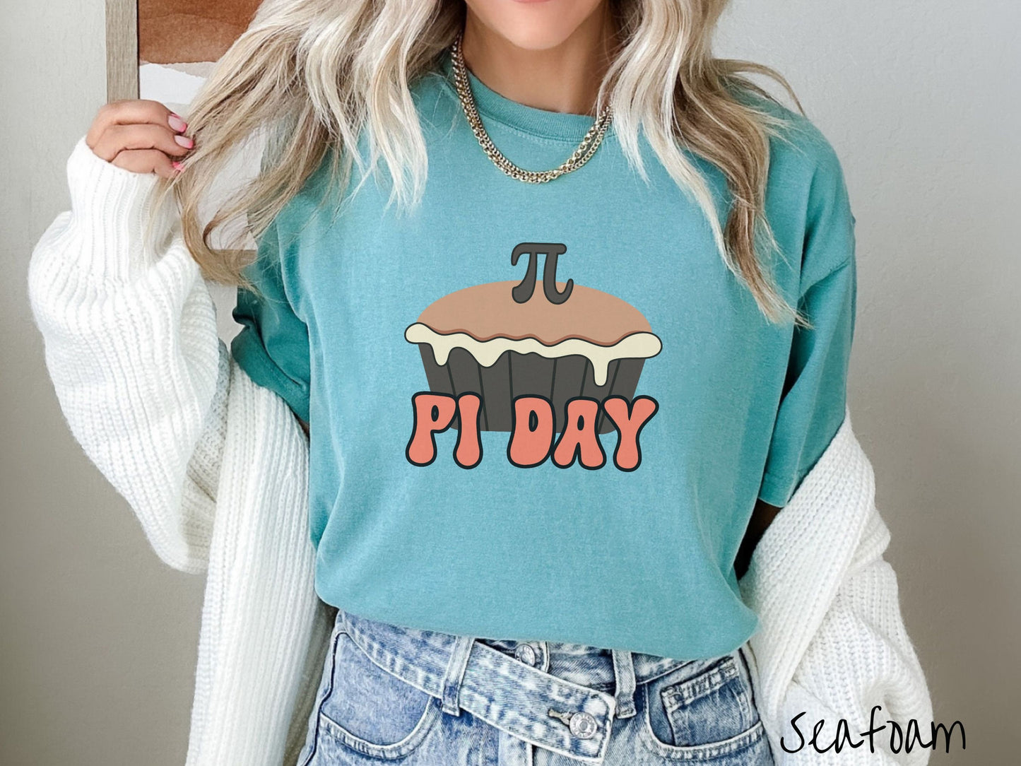 A woman wearing a cute, vintage seafoam colored comfort colors shirt with the text Pi Day in orange font, above that is a brown pumpkin pie with a pi symbol on the top of the pie.