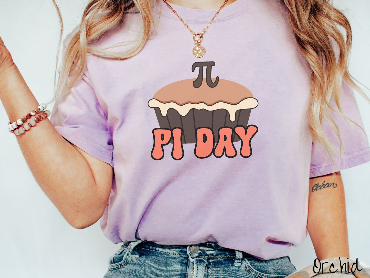 A woman wearing a cute, vintage orchid colored comfort colors shirt with the text Pi Day in orange font, above that is a brown pumpkin pie with a pi symbol on the top of the pie.