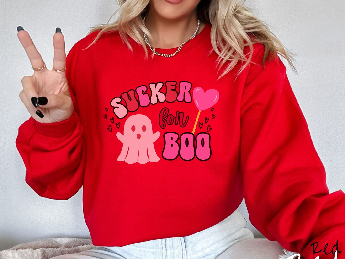 A woman wearing a cute red colored sweatshirt with the text saying Sucker for Boo, with a pink ghost with its arms open, a lollipop with a pink heart, and hearts floating in the air.