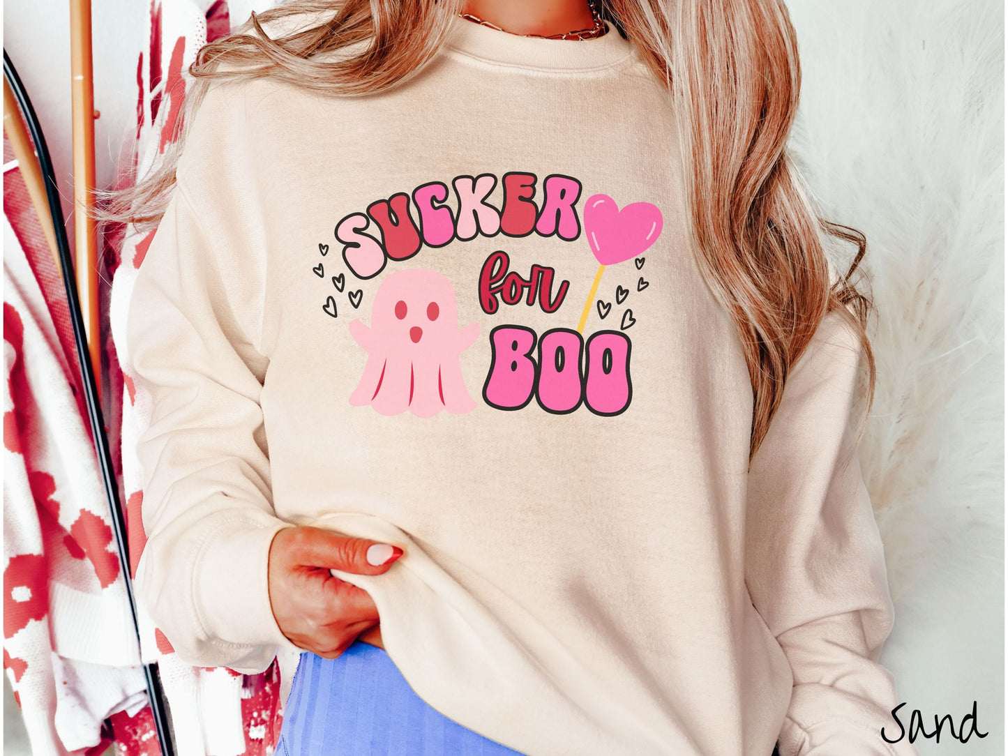 A woman wearing a cute sand colored sweatshirt with the text saying Sucker for Boo, with a pink ghost with its arms open, a lollipop with a pink heart, and hearts floating in the air.