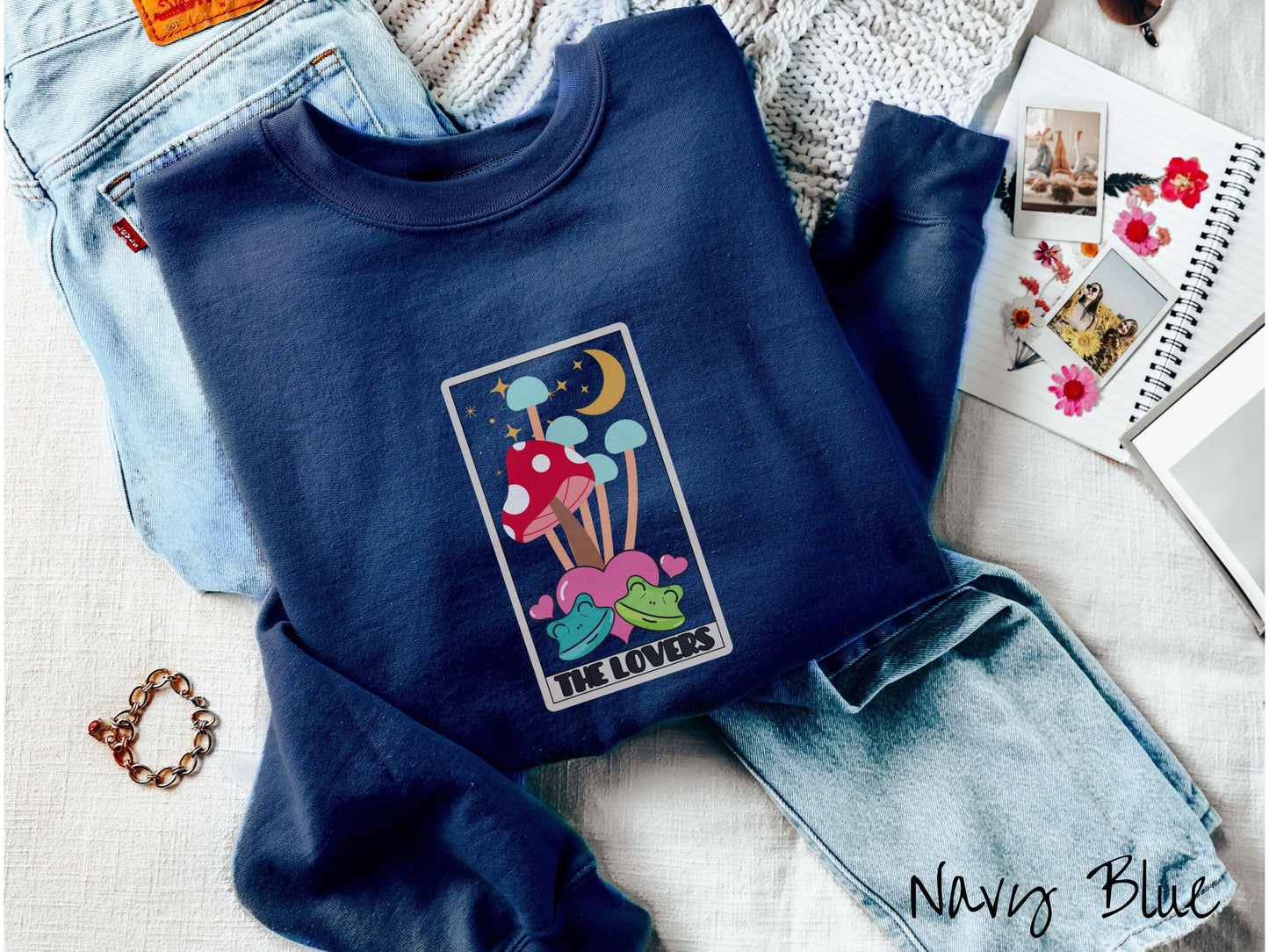 A navy blue colored sweatshirt with a large red mushroom and 4 turquoise mushrooms in the background, all under a crescent yellow moon and yellow stars, two green frogs smiling in front of pink hearts below the mushrooms.