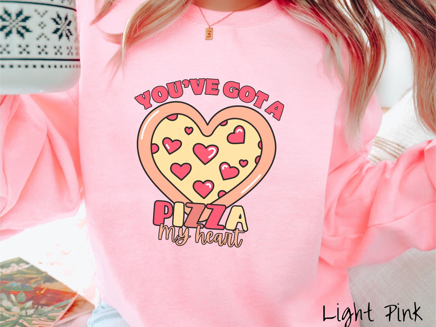 A woman wearing a cute light pink colored sweatshirt with red text Youve Got a Pizza My Heart around a heart-shaped pizza with heart-shaped red pepperonis.