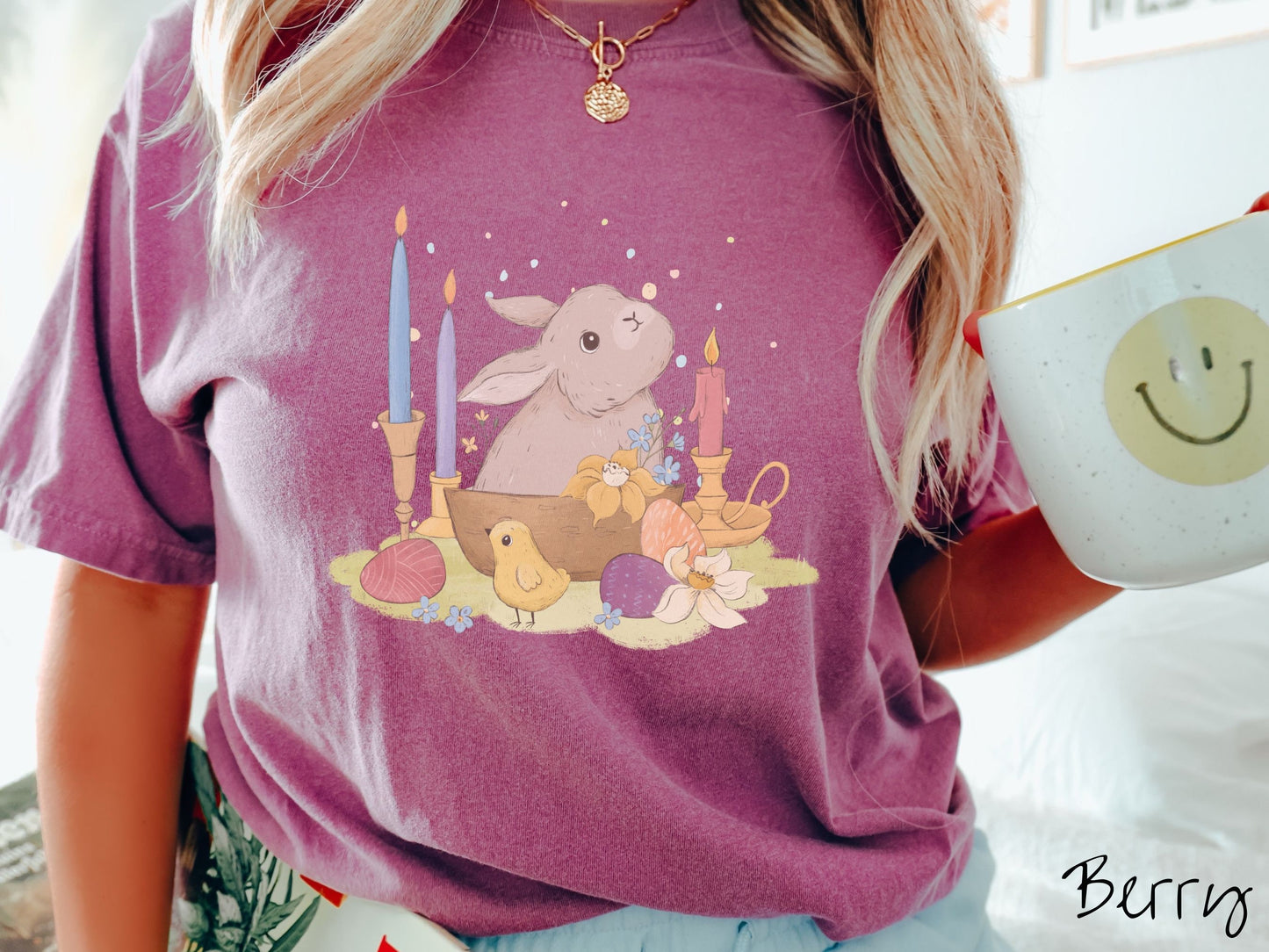 A woman wearing a cute, vintage berry colored shirt with a gray bunny rabbit sitting in between three lit candles, some red, orange, and purple eggs, and yellow and white flowers.