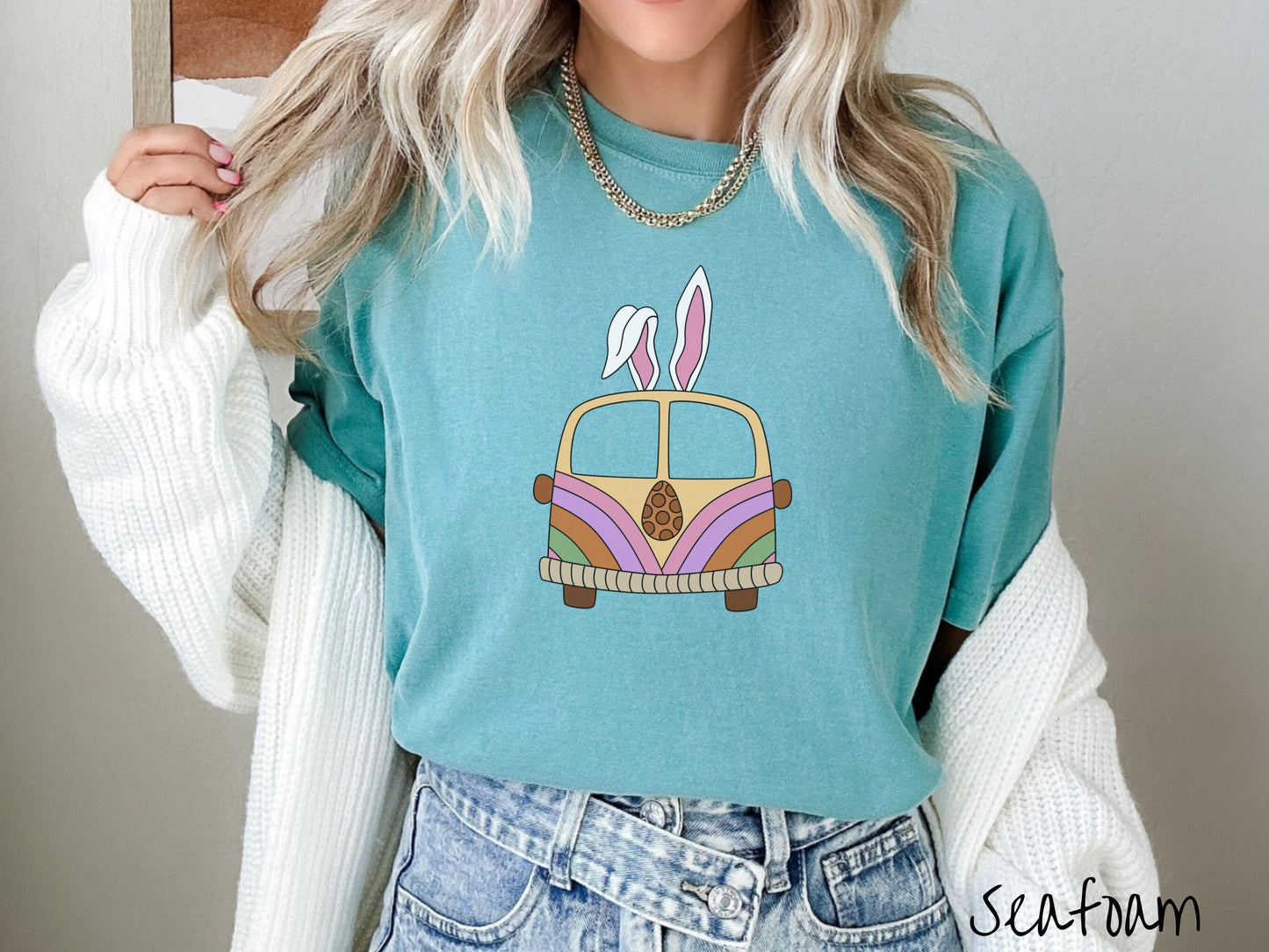 A woman wearing a cute, vintage seafoam colored shirt with an old-fashioned, hippie style van with pastel-colored stripes on the front and light orange paint covering the rest. The van has a brown egg on the front and two, floppy bunny ears on top.