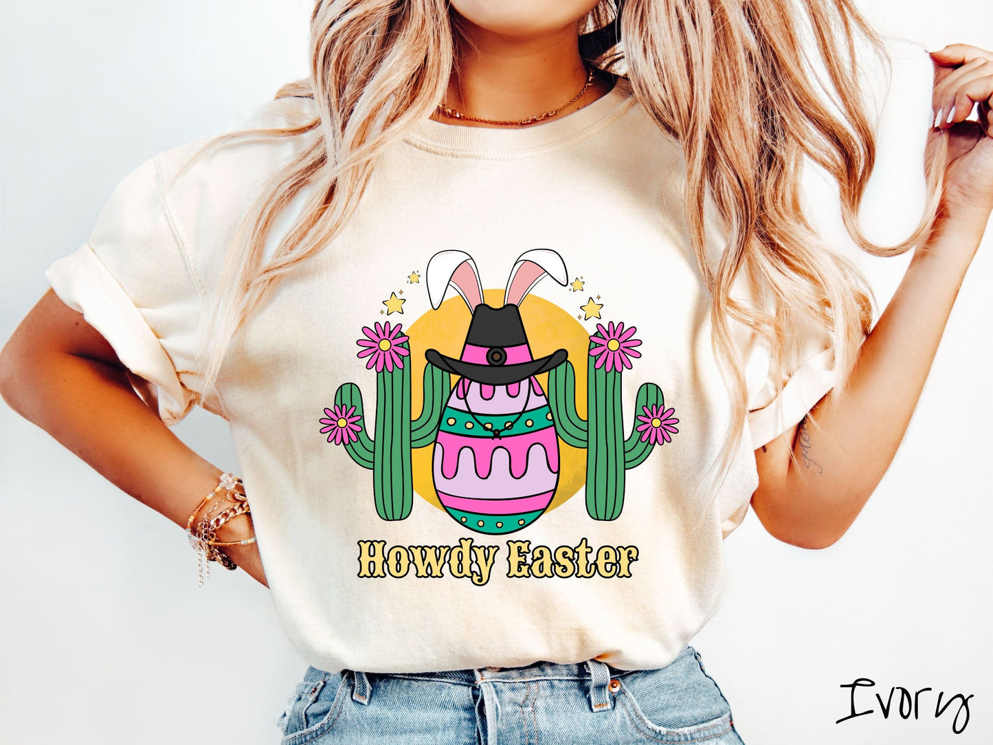 A woman wearing a cute, vintage ivory colored shirt with an Easter egg with green, pink and purple stripes, wearing a black cowboy hat and white bunny ears, and it’s standing in between two green cacti with pink flowers in front of the sun.