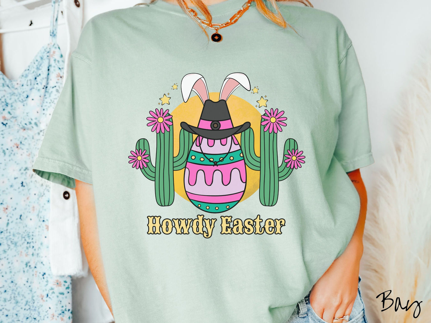 A woman wearing a cute, vintage bay colored shirt with an Easter egg with green, pink and purple stripes, wearing a black cowboy hat and white bunny ears, and it’s standing in between two green cacti with pink flowers in front of the sun.