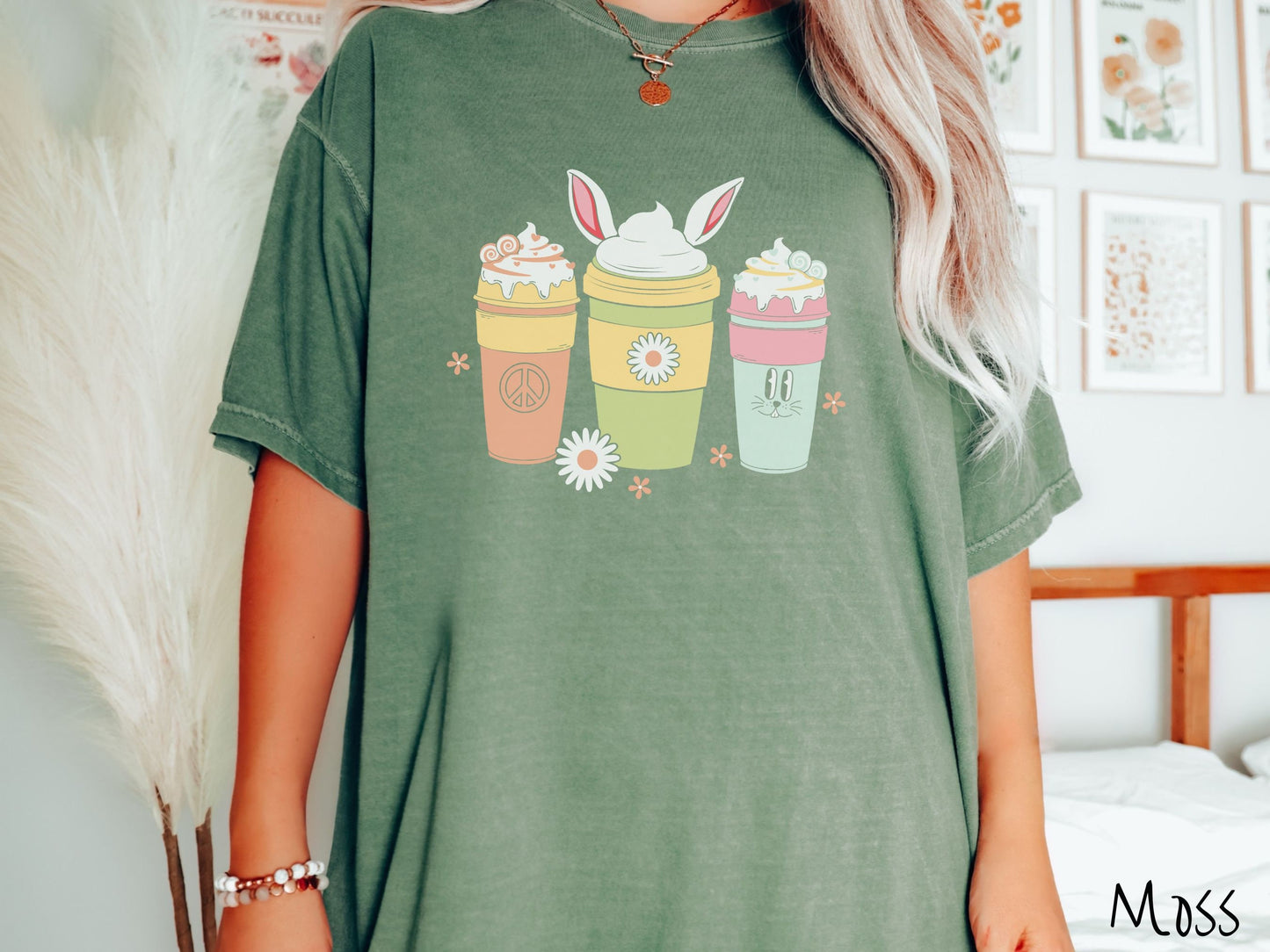 A woman wearing a cute, vintage moss colored shirt with three coffee cups on the front. One is orange with whipped cream and a peace sign. The middle is green with whipped cream, and bunny ears, and the third is light blue with whipped cream.