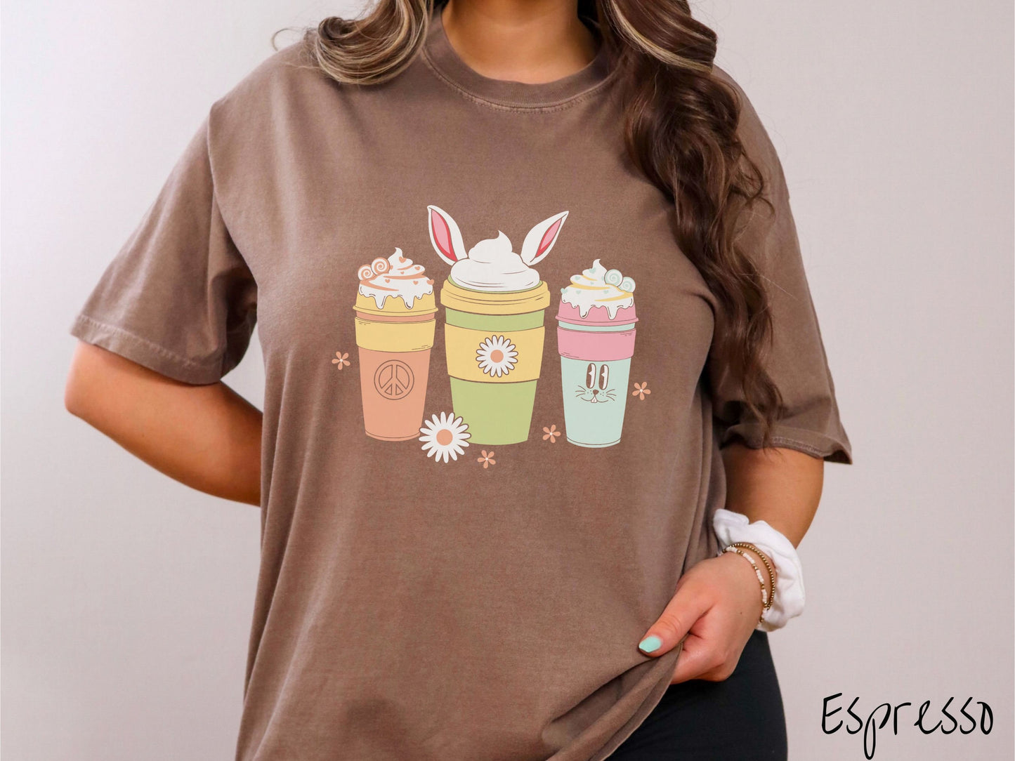 A woman wearing a cute, vintage espresso colored shirt with three coffee cups on the front. One is orange with whipped cream and a peace sign. The middle is green with whipped cream, and bunny ears, and the third is light blue with whipped cream.