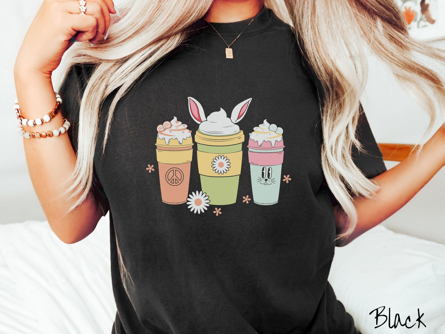 A woman wearing a cute, vintage black colored shirt with three coffee cups on the front. One is orange with whipped cream and a peace sign. The middle is green with whipped cream, and bunny ears, and the third is light blue with whipped cream.
