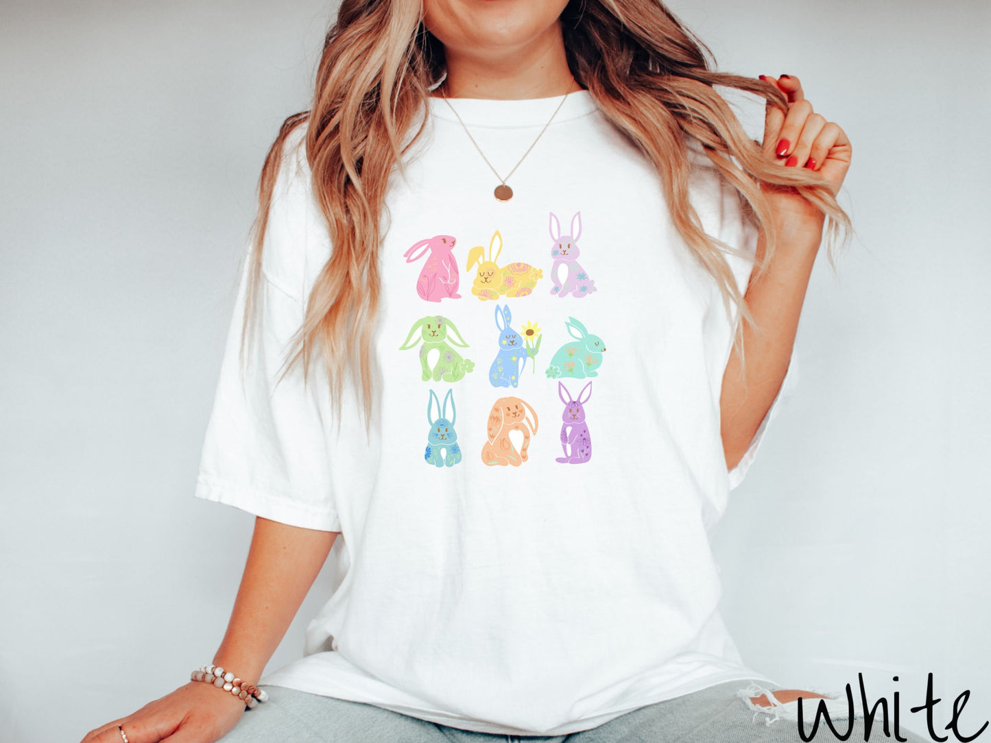 A woman wearing a cute, vintage white colored shirt with a three by three grid of colorful bunny rabbits. They are left to right pink, yellow, purple, green, blue, seafoam, light blue, orange, and purple and have flowers and plants drawn on them.