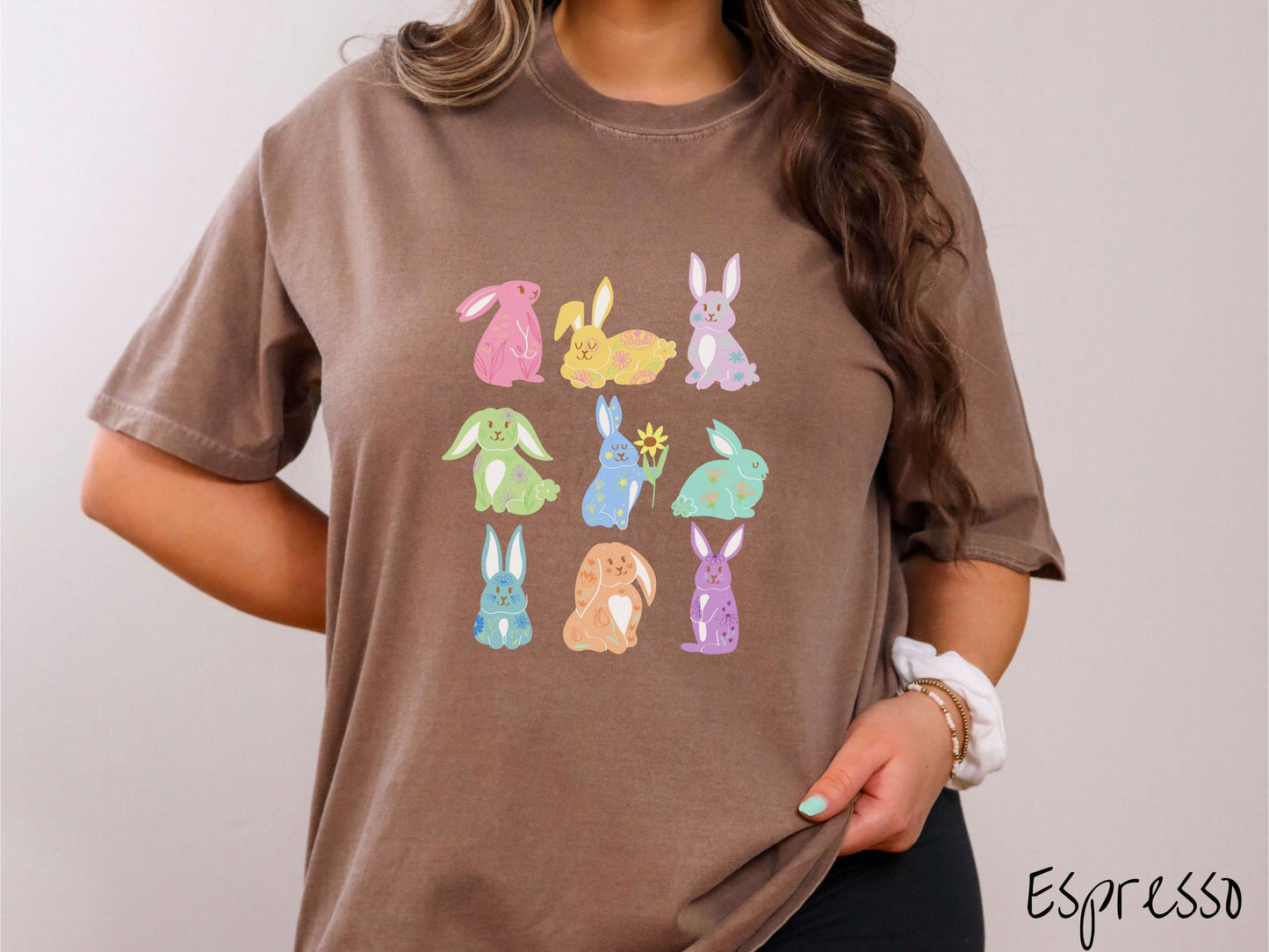 A woman wearing a cute, vintage espresso colored shirt with a three by three grid of colorful bunny rabbits. They are left to right pink, yellow, purple, green, blue, seafoam, light blue, orange, and purple and have flowers and plants drawn on them.