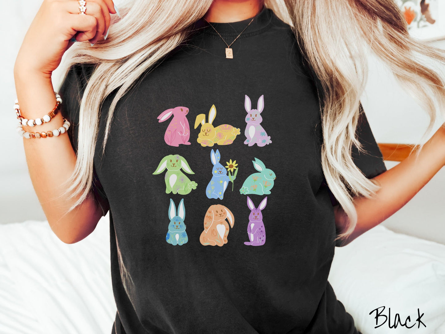 A woman wearing a cute, vintage black colored shirt with a three by three grid of colorful bunny rabbits. They are left to right pink, yellow, purple, green, blue, seafoam, light blue, orange, and purple and have flowers and plants drawn on them.