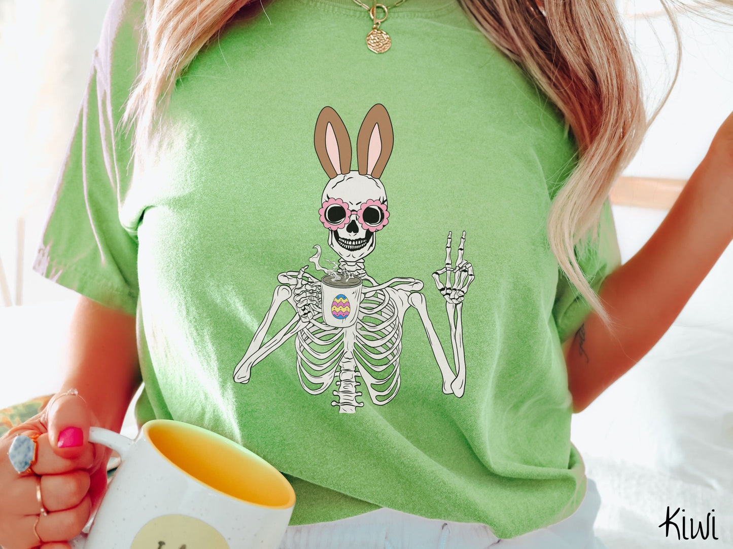 A woman wearing a cute, vintage kiwi colored shirt with a pink-toned skeleton wearing pink glasses and brown bunny ears, holding a steamy cup of coffee while holding up a peace sign with the other hand. The white coffee cup has a colorful Easter egg
