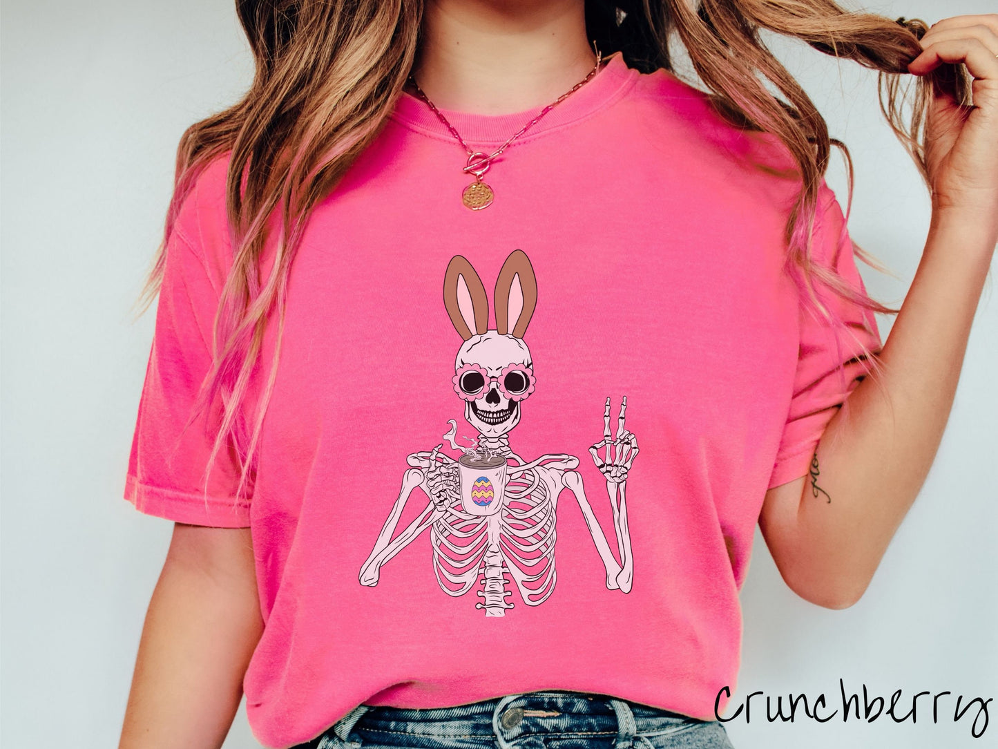 A woman wearing a cute, vintage crunchberry colored shirt with a pink-toned skeleton wearing pink glasses and brown bunny ears, holding a coffee cup and holding up a peace sign with the other hand. The white coffee cup has a colorful Easter egg