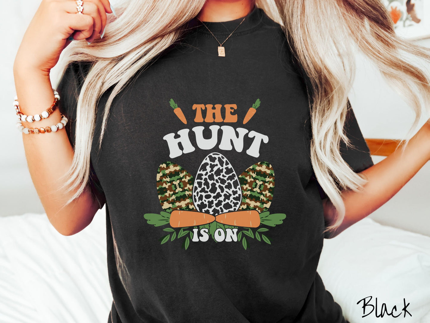 A woman wearing a cute, vintage black colored shirt with the text The Hunt Is On in orange and white font, with three Easter eggs painted in woodland camo and black and white camo in front. Above and below the eggs are carrots with leafy ends.