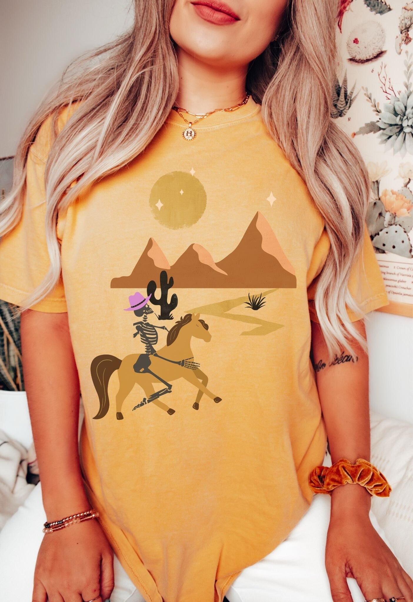A woman wearing a vintage, mustard colored shirt with a skeleton cowgirl wearing a pink cowboy hat riding a brown horse through the eset at night, cacti and three mountains in the background with a full moon and stars in the sky.