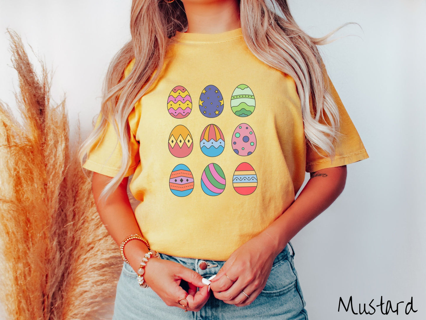 A woman wearing a cute, vintage mustard colored shirt with a three by three grid of colorful Easter eggs. One is pink, red, and yellow, another blue with stars, and another is different shades of green next to five more egg designs.