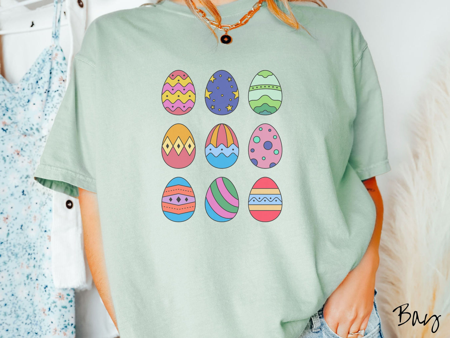 A woman wearing a cute, vintage bay colored shirt with a three by three grid of colorful Easter eggs. One is pink, red, and yellow, another blue with stars, and another is different shades of green next to five more egg designs.